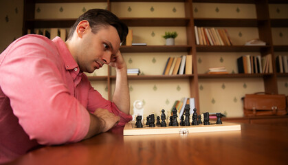 A person in a pink shirt thoughtfully looks at a chessboard, resting his chin on his hand. The scene is set in a cozy indoor space with bookshelves and a bag in the background.