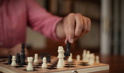 A person in a pink shirt moves a black queen on a chessboard, carefully positioning it over white pieces. The scene is set indoors with a warm atmosphere and a blurred bookshelf background.