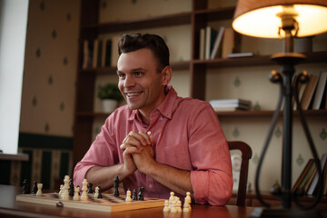 A person in pink shirt smiles while playing chess,with hands clasped in front of the board. The scene is set in cozy indoor space with bookshelves,plants,and warm lighting, creating relaxed atmosphere