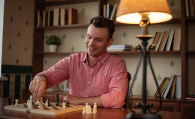 A person in a pink shirt smiles while playing chess, moving a white piece on the board. The cozy...
