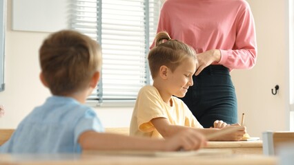 Fototapeta premium Beautiful teacher explaining test while caucasian boy doing classwork and listening explanation. Cute child looking at teacher while taking a notes or writing answer in paper at classroom. Pedagogy.