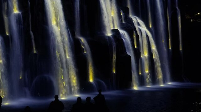 Visitors gather at a city park to admire a stunning illuminated waterfall display. The colorful lights enhance the beauty of cascading water under the nighttime sky
