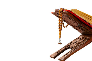 A close-up shot of an open Quran on a traditional wooden stand (rahle) with a string of prayer beads (tesbih) laid on top. The image is set against a plain gray background, highlighting the Quran and 