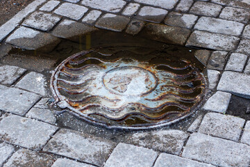 A manhole cover sits on a brick sidewalk by a puddle