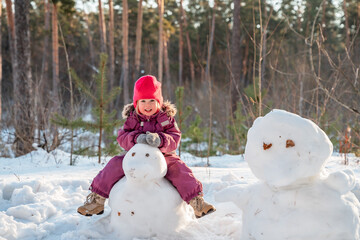 Cute little Caucasian child toddler girl sculpting a snowman in a snowy forest. Winter activities...