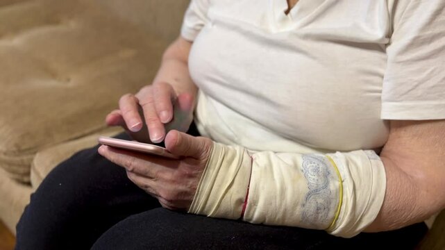 Elderly woman with broken arm width cast for the arm sitting at beige couch holding smartphone in her hand looking for important information using rose smartphone