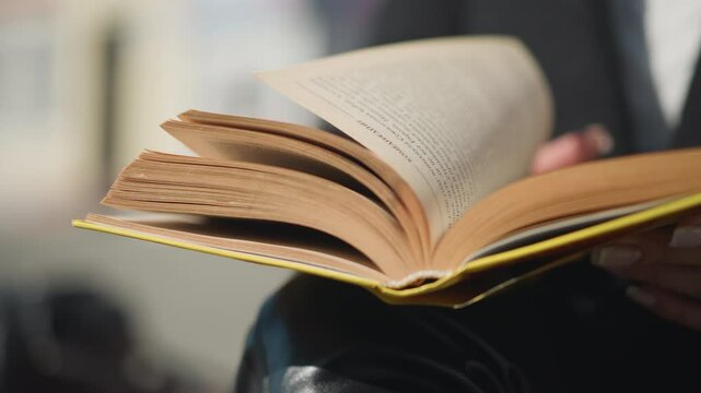 Close-up of book pages being flipped by a hand adorned with a ring and polished nails, dressed in black clothing, the background is blurred