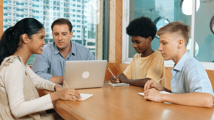 Smart girl ask teacher a question while show idea while diverse student working classwork. Group of multicultural children pointing at notebook while happy boy sharing idea at classroom. Edification.