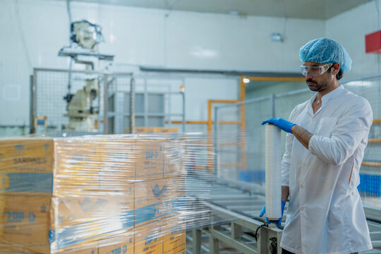Factory worker in protective clothing wrapping pallets with plastic film, ensuring safe packaging in a modern warehouse for food production and distribution.