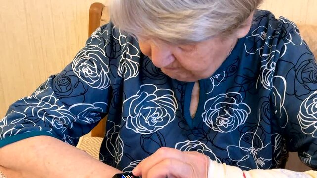 The black smart band with display touchpad and heart rate monitor. The old woman with broken arm with cast for the arm sitting at sofa touches and looks at the wristband of pulse monitor indoors