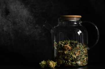 A glass teapot is filled with dried herbs. The pot stands in front of a dark background. There is...