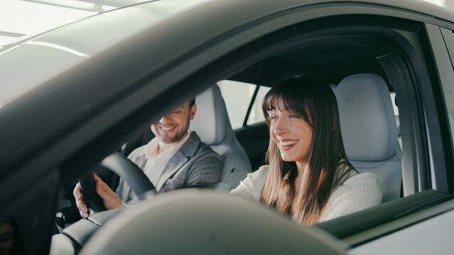 Side camera view of positive woman exclaiming with joy while sitting at driver seat in new car. Husband smiling while sitting besides her. Shopping for new vehicle. Positive emotions.