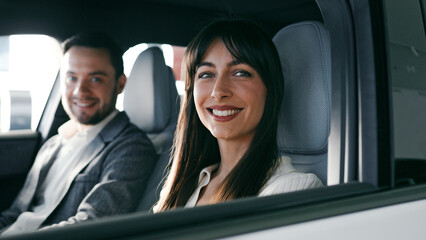 Side view of charming Caucasian couple sitting inside of new vehicle. Female trying out steering wheel. Imagining driving while listening to husband talking. Looking at camera and smiling.