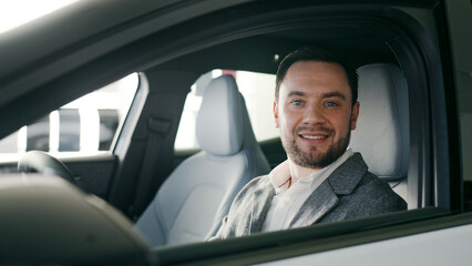 Bearded Caucasian male sitting in car. Looking at dashboard and trying quality of materials. Smiling to camera while looking directly. Shopping for new vehicle in dealership. Wearing white shirt.