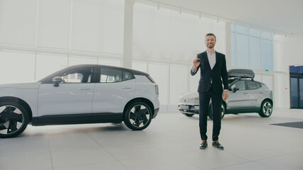 Stylish Caucasian male in black suit standing in middle of car dealership. Showing to camera keys...