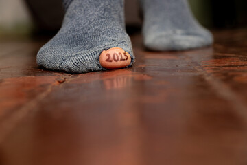 2025 written on a toe and the toe sticks out of a hole in a torn sock on the floor, 2025