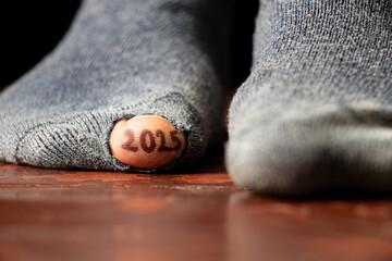 2025 written on a toe and the toe sticks out of a hole in a torn sock on the floor, 2025
