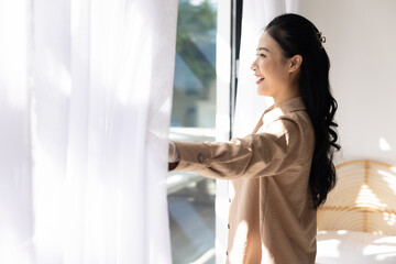 Cheerful young Asian hotel guest girl drawing white transparent curtains in morning, looking at...