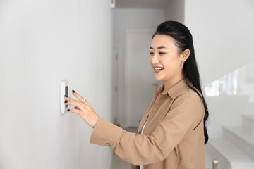 Happy young Asian homeowner woman typing on wall mounted climate control panel, regulating...