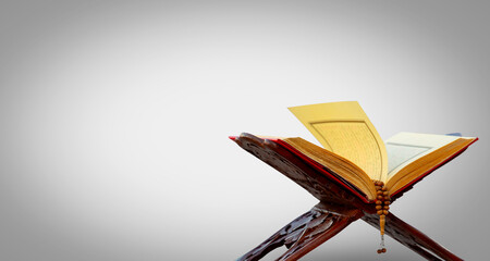 A close-up shot of an open Quran on a traditional wooden stand (rahle) with a string of prayer beads laid on top. The image is set against a plain gray background, highlighting the Quran and prayer