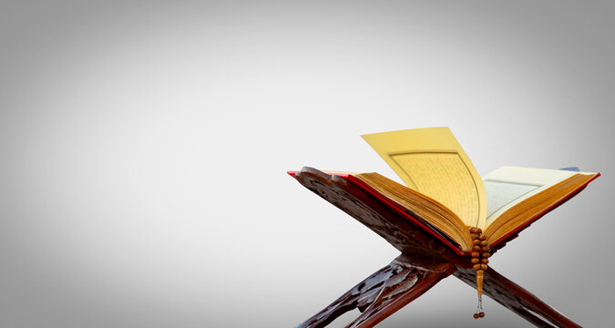 A close-up shot of an open Quran on a traditional wooden stand (rahle) with a string of prayer beads laid on top. The image is set against a plain gray background, highlighting the Quran and prayer