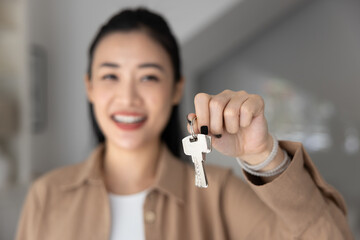 Happy new tenant woman holding hanging key from new real estate property, smiling at camera in blurred background, enjoying being at new home. Young adult Asian girl getting flat for rent