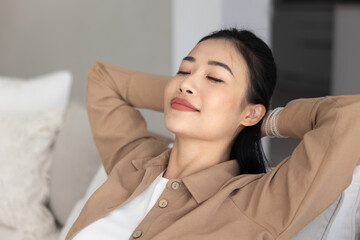 Serene positive beautiful Asian woman resting on comfortable sofa, leaning on soft back with closed...