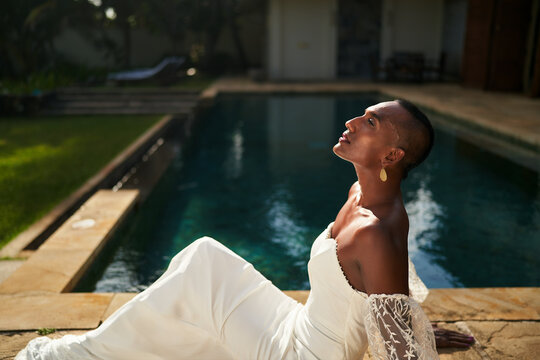 Bride in white dress by pool on sunny day. Gender fluid person enjoys wedding at villa. LGBTQ celebration, modern love scene. Beautiful garden, serene water. Diverse romance. - Powered by Adobe