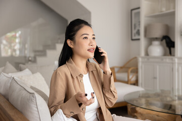 Serious young Asian cellphone user girl speaking on mobile phone at home, sitting on cozy couch,...