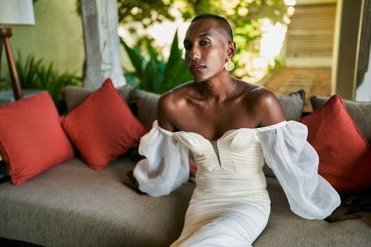 Ambiguous person in elegant white dress sitting at villa. Gender fluid bride on couch, trees in background. Unique LGBTQ wedding theme. Fashionable, stylish, diverse, inclusive imagery.