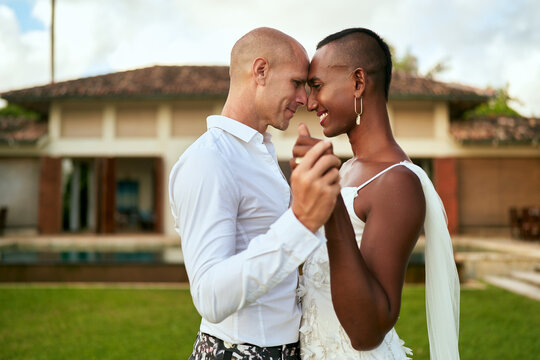 LGBTQ couple dances in villa wedding. Bride in white dress, groom in bold suit. Tropical background creates romantic atmosphere. Joyful celebration scene, inclusive love.