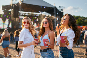 Group of happy young women with sunglasses enjoy summer music festival on sandy beach. Holding...
