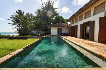 Sunny tropical villa with pool, palm trees by ocean. Vintage architecture, wooden shutters open. Relaxing holiday vibe in exotic paradise. Poolside chairs invite leisure. Charming getaway spot.