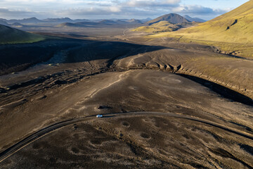 Aerial perspective of a car navigating a sparse basalt road Landmannalaugar in Iceland, surrounded by barren landscapes and bathed in warm sunlight