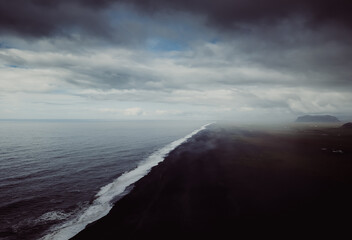 A dramatic aerial composition showcasing a black sand beach Reynisfjara stretching endlessly with moody clouds and tranquil ocean waters. A breathtaking depiction of nature's raw beauty in Iceland