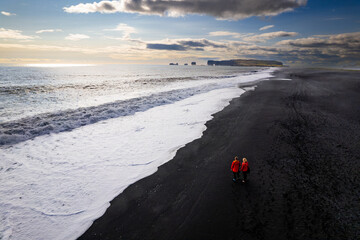 Aerail view couple walking along a stunning black sandy beach Reynisfjara , sunset in Iceland VIK Myrdal in Golden Circle area with Atlantic Ocean waves crashing and volcanic rock formations
