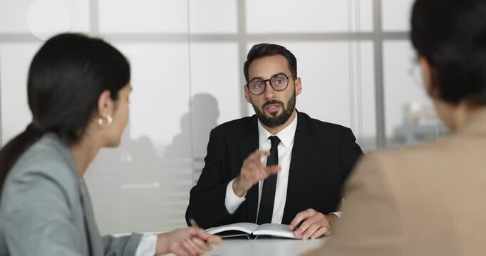 Hispanic businessman in black suit presenting information, explain point to women colleagues during briefing or meeting in boardroom. Team engaged in focused discussion seated across conference table