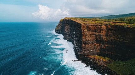 A stunning aerial view of a rugged cliff meeting the ocean, with waves crashing against the rock face under a cloudy sky.