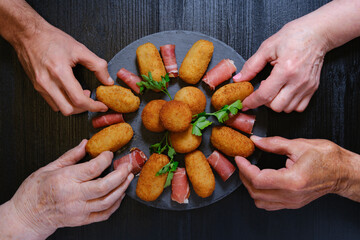 Hands approaching a slate platter to pick up croquettes and slices of Serrano ham