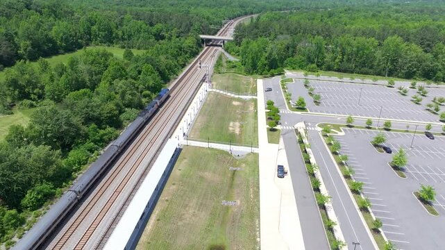 Daytime overhead view from a drone of two parallel railroad train tracks and a commuter passenger train near fields and a commuter parking lot.