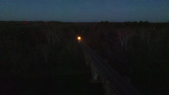 Nightime evening aerial view from a drone of two parallel railroad train tracks and a passenger train crossing a river on an elkevated bridge and arriving at a railway station.