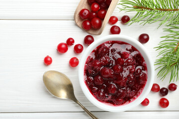 Tasty cranberry sauce in bowl, berries, spoon and fir tree branches on white wooden table, top view