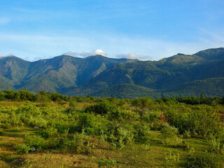 A full-length outdoor photograph of a landscape with various mountain ranges under a clear, light blue sky. The overall look conveys the peaceful, natural feel of a mountainous region.