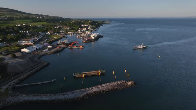 Lough Foyle Ferry, County Donegal, Ireland, June 2023. Drone descends slowly towards a transport vessel approaching Greencastle Harbour filled with boats and heavy machinery in the soft evening light.