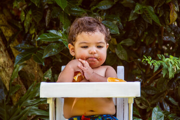 9-month-old baby eating orange in his white highchair, outdoors.