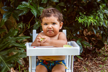 9-month-old baby eating lemon in his white highchair, outdoors.