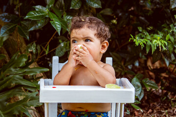 9-month-old baby eating lemon in his white highchair, outdoors.