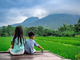 Back view of small Asian children relaxing sitting on bamboo litter in a green rice field with a sky of mountains and clouds.