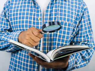 Cropped adult Asian man searching something inside a book using magnifying glass. Selective focus