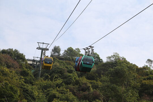 The cable car on the mountain is green, yellow, and blue in color, with two cars hanging from it, flying high above the trees below